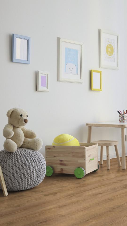 Kinderzimmer mit Holzboden, Kindertisch, Teddybär, Spielzeugkiste und großem Fenster mit weißen Vorhängen.