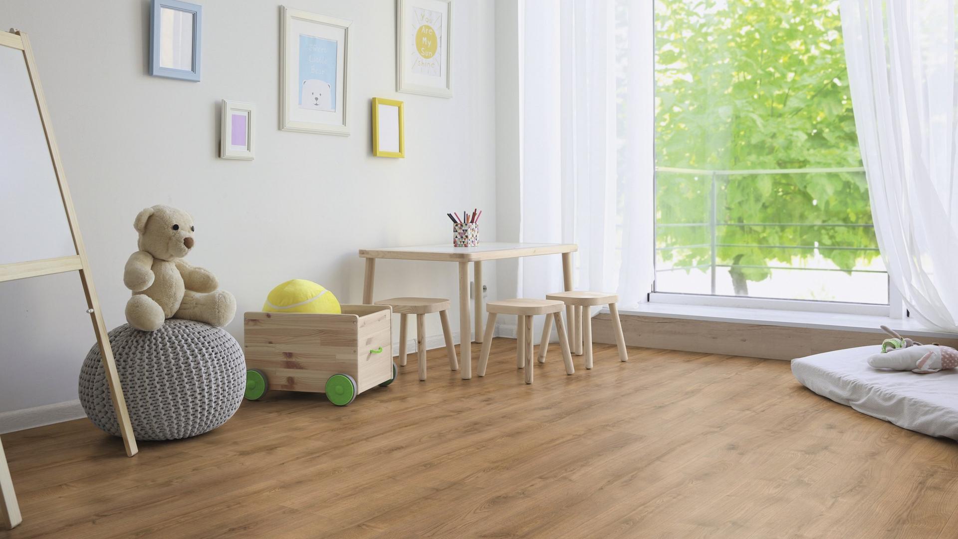 Cameretta con pavimento in legno, tavolo, orsacchiotto, scatola dei giochi e finestra grande con tende bianche.